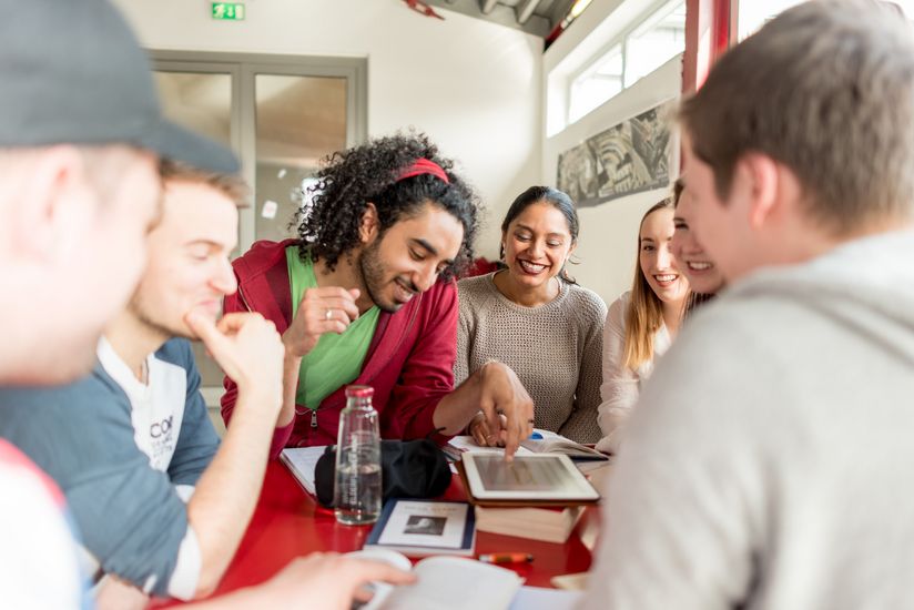 Studierende Eine Gruppe von Studierenden sitzt an einem Tisch. Sie lachen und blicken teilweise auf ein Tablet vor ihnen auf dem Tisch.