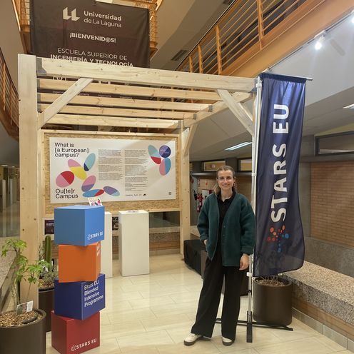 Janina Ebner STARS EU Projekt Outer Campus Junge Frau steht vor einem Pavillon aus Holz in einem Gebäude. Rechts neben ihr steht eine Fahne mit dem Schriftzug STARS EU. Lonks neben ihr stehen bunte Würfel.