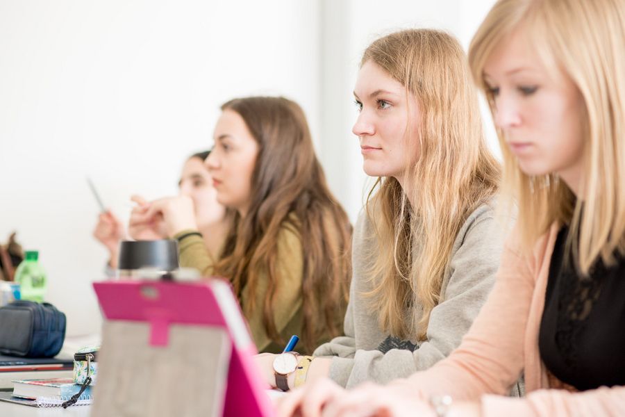 Studierende in Vorlesung Vier Studentinnen sitzen an einer Tischreihe lauschen der Vorlesung und schreiben mit.