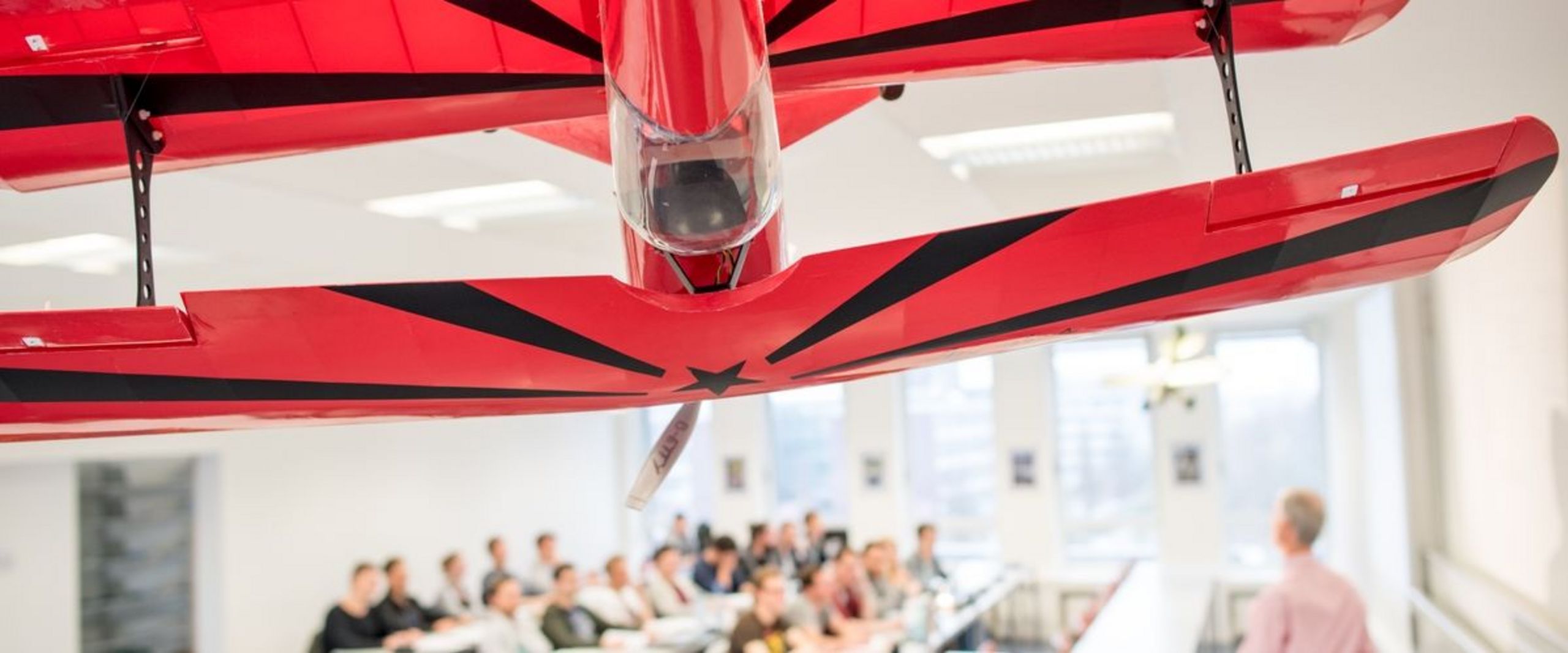Auf dem Bild ist ein großes rotes Modellflugzeug zu sehen, das von der Decke eine Vorlesungssaales hängt. Im Hintergrund ist eine Grube Studierender und ein Lehrender im Seminarraum zu sehen.