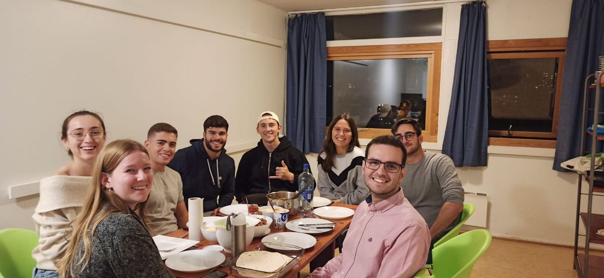 Auf diesem Bild sieht man eine Gruppe von acht Studierenden gemeinsam an einem Tisch sitzen. Sie haben einige Töpfe mit gemeinsam gekochten Essen auf dem Tisch vor sich stehen und freuen sichauf ihr Abendessen. 