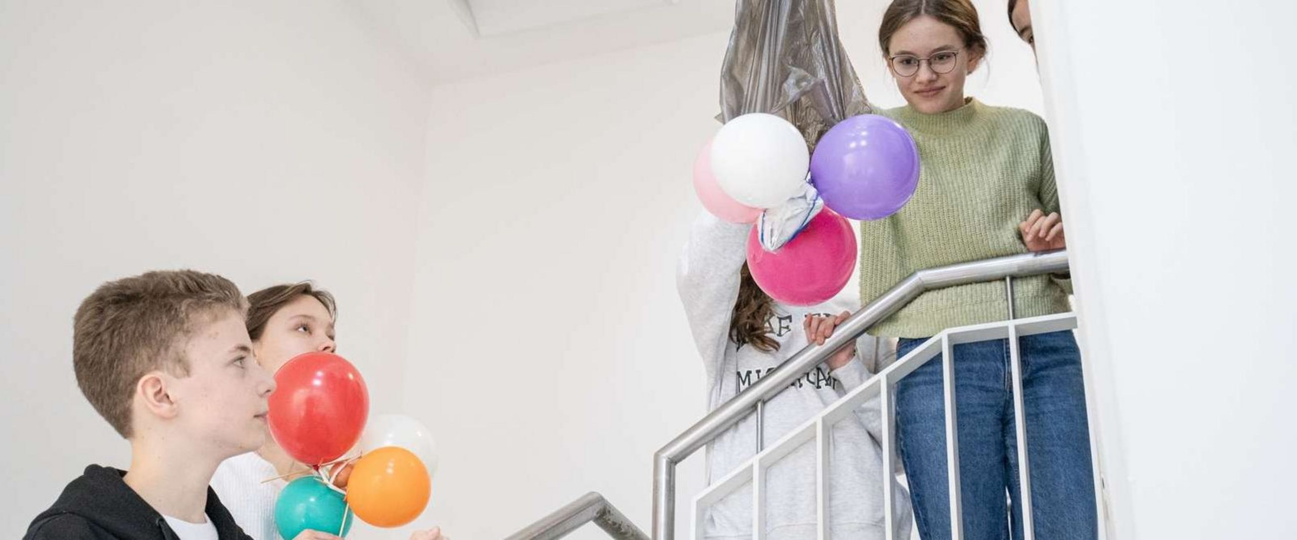 Eine Schülerin hält eine Konstruktion aus Müllbeutel und Luftballons rund um ein rohes Ei in die Luft, bereit, diese im Treppenhaus aus einem höheren Stockwerk fallen zu lassen. Andere Schüler:innen schauen gespannt zu. 