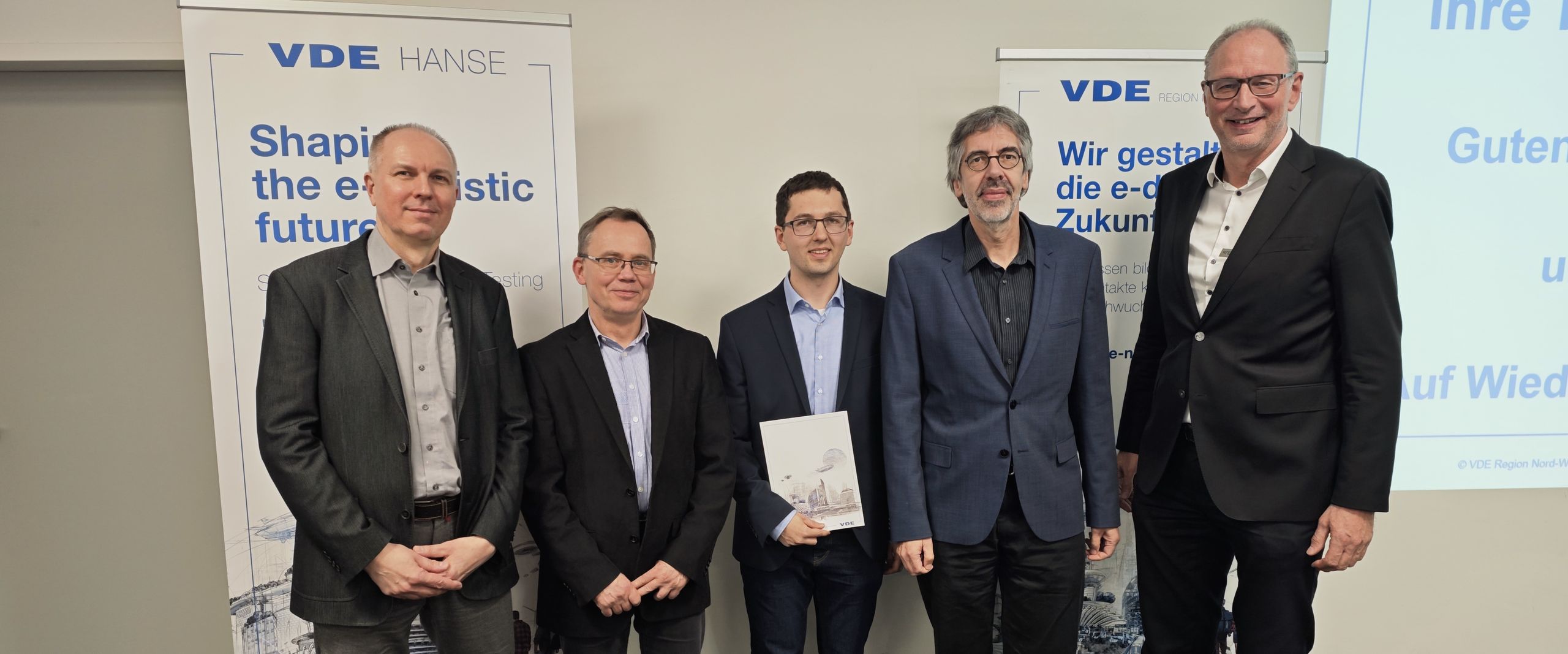 Gruppenbild mit Männern im Anzug, die vor Roll-ups stehen. Einer in der Mitte hält eine Urkunde.