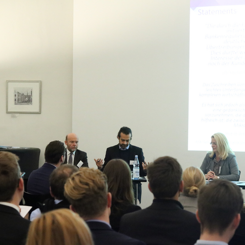 M&auml;nne rund Frauen sitzen in einem Raum und diskutieren. Hinter ihnen ist eine Powerpointpr&auml;sentation.