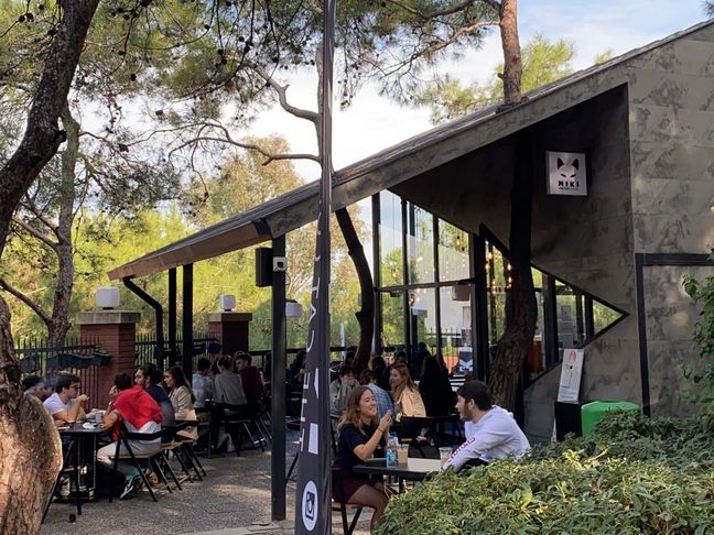 Restaurant im Campuspark der Izmir University of Economics Das Bild zeigt die Außenterasse eines Restaurants im Campuspark der IUE in Izmir. Alle Plätze sind besetzt und die Studierenden unterhalten sich angeregt. Rund um das Restaurant stehen Pinien.