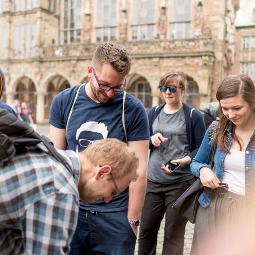 HSB Studierende l&auml;chelnd nach unten blickend im Kreis stehend mittig im Kreis ein weiterer HSB Studierender zeigend auf den Boden. 