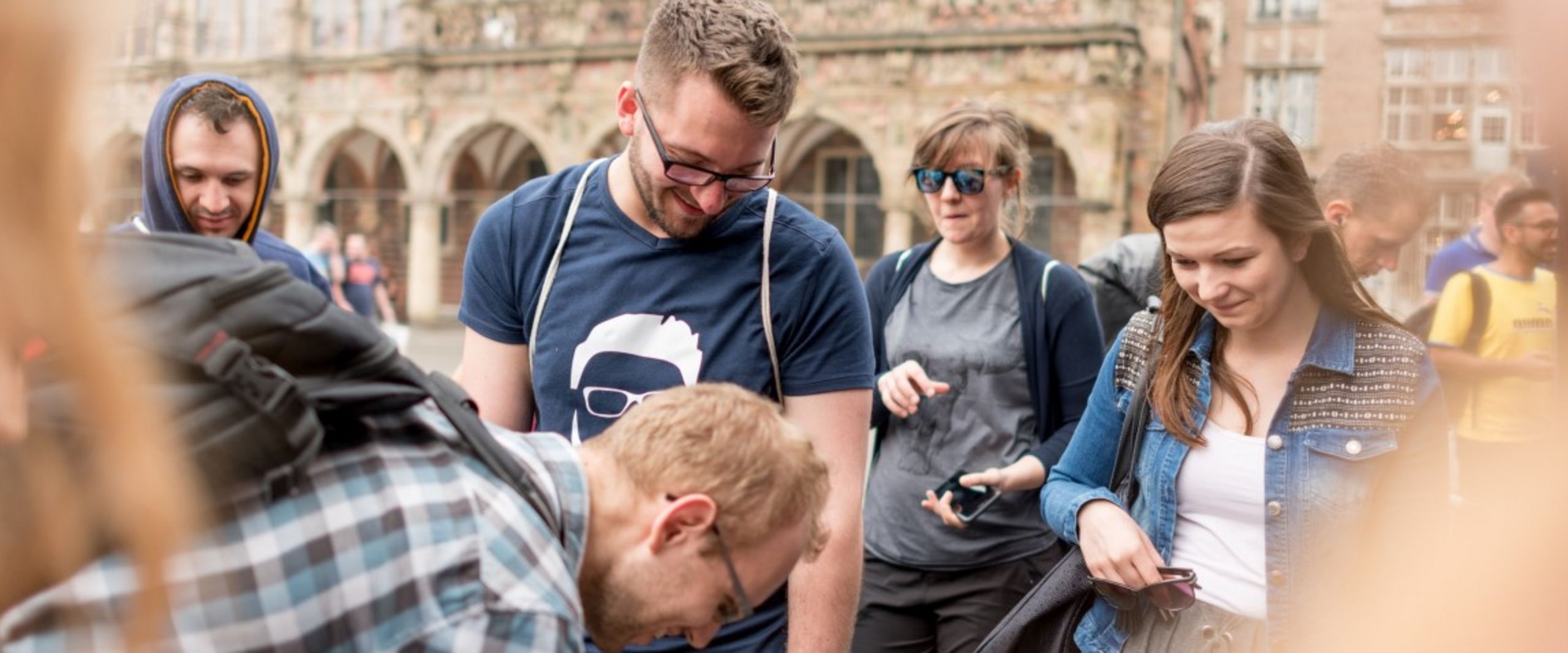 HSB Studierende l&auml;chelnd nach unten blickend im Kreis stehend mittig im Kreis ein weiterer HSB Studierender zeigend auf den Boden. 