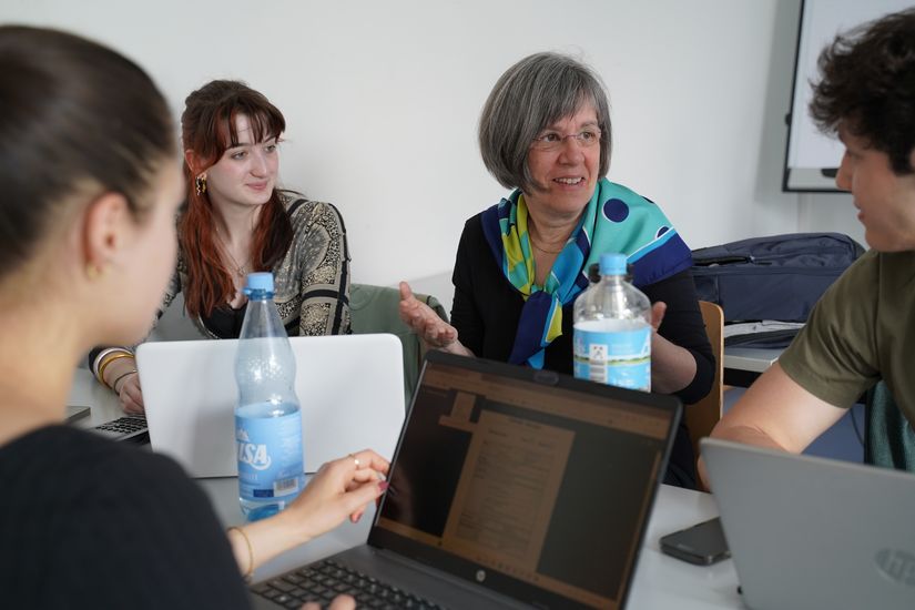 Studierende und Dozentin arbeiten zusammen am Tisch. Vor ihnen stehen Laptops.
