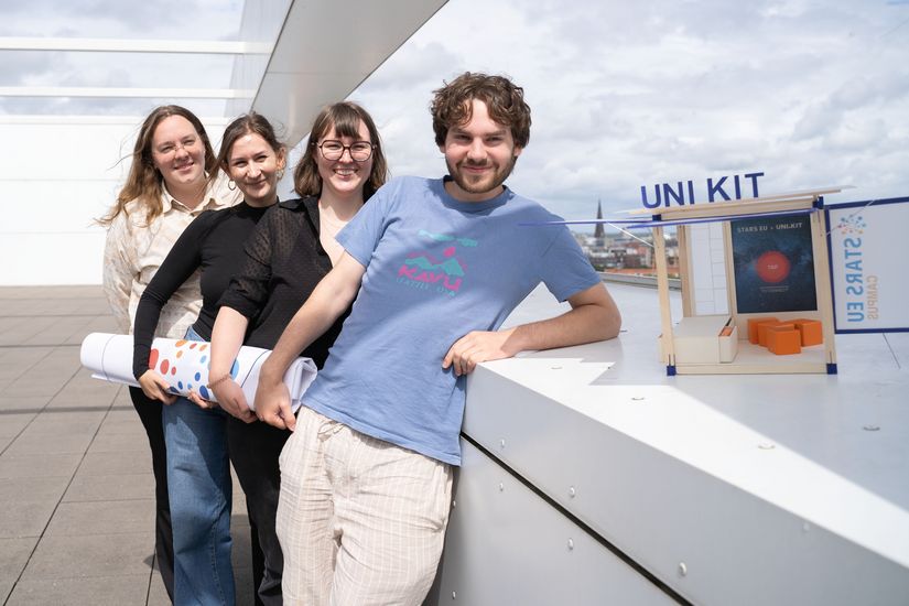 Ein junger Mann und drei junge Frauen stehen hintereinander und lachen in die Kamera. Neben dem Mann steht ein kleines Architekturmodell aus Holz.