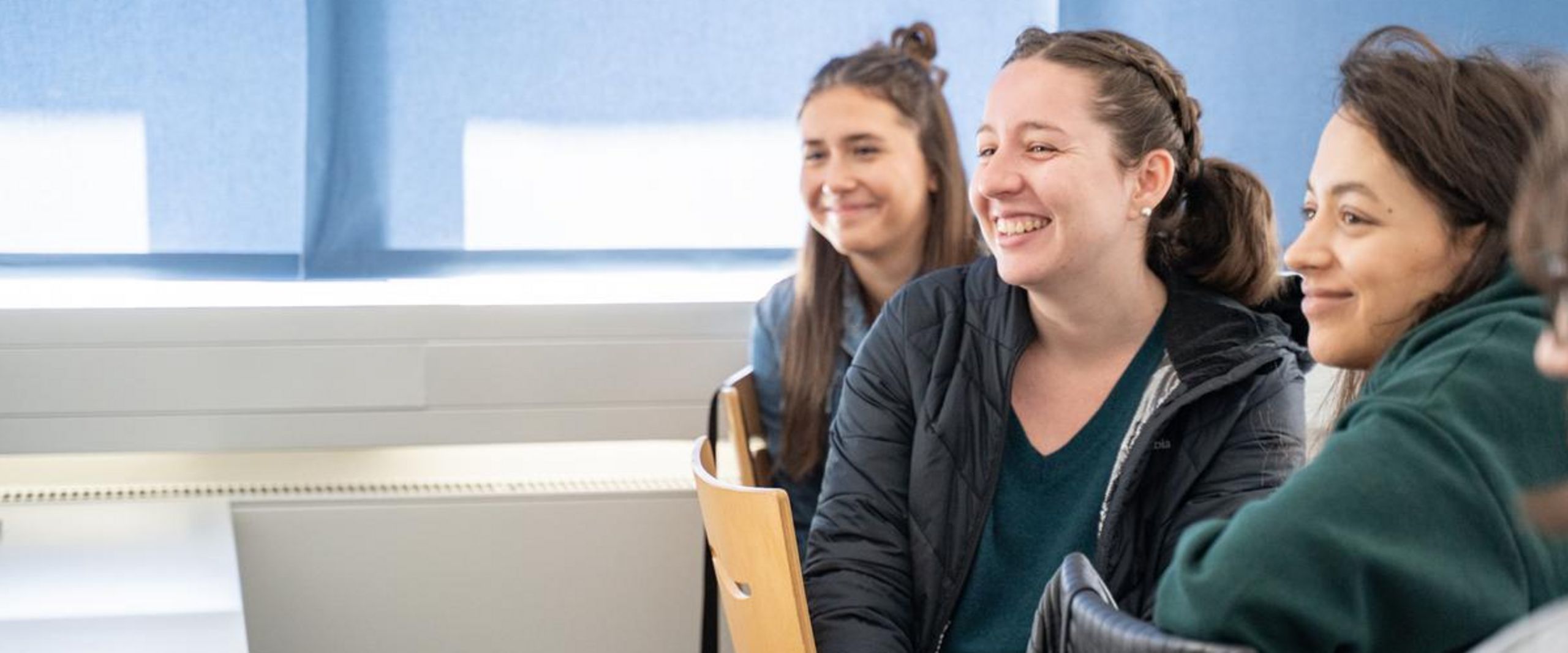 Auf dem Bild sind drei Studentinnen zusehen, die in einer Reihe an einem Tisch sitzen und sich jeweils nach hinten umdrehen. Dort scheint etwas lustiges zu passieren. 