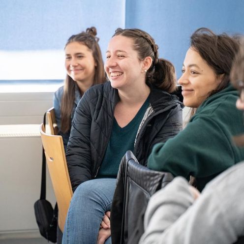 Auf dem Bild sind drei Studentinnen zusehen, die in einer Reihe an einem Tisch sitzen und sich jeweils nach hinten umdrehen. Dort scheint etwas lustiges zu passieren. 