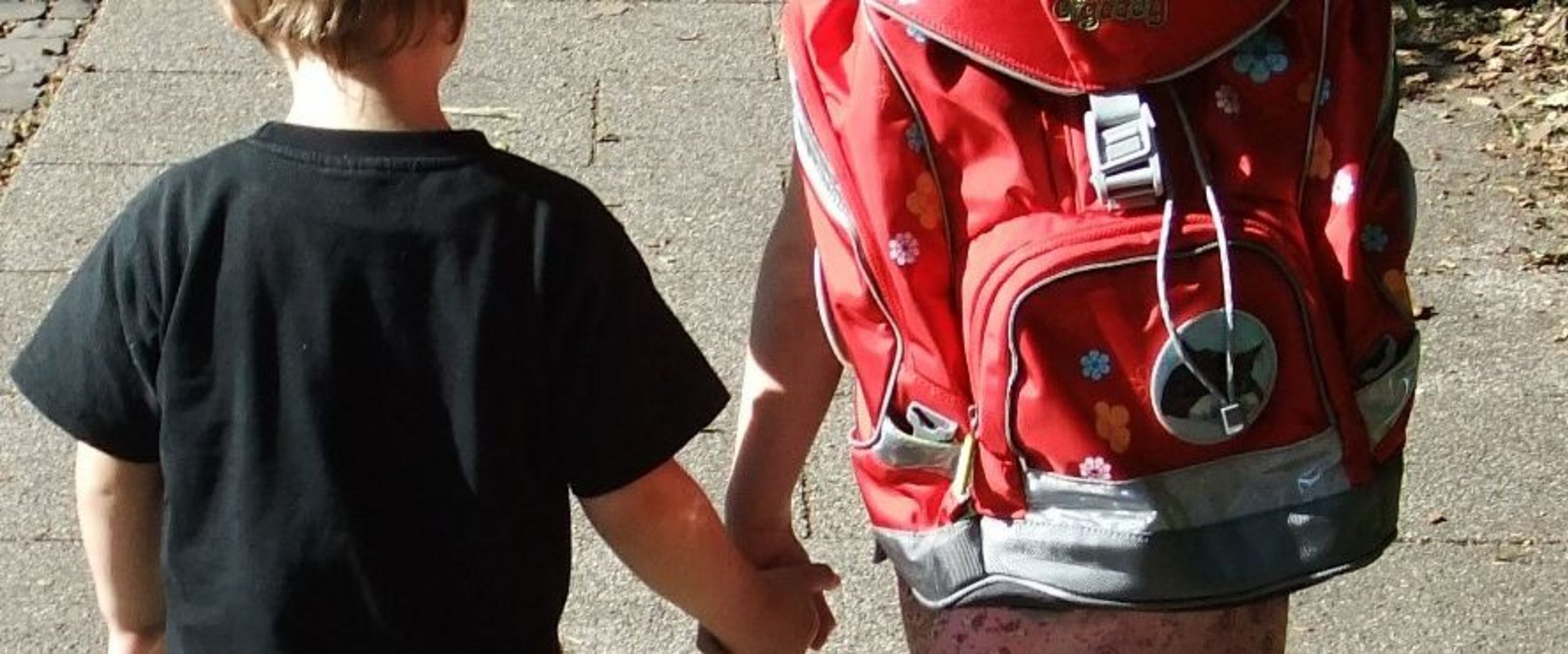 Bildausschnitt von zwei Kinder (Ansicht von hinten), die sich an der hand halten. das rechte trägt einen Schulranzen.