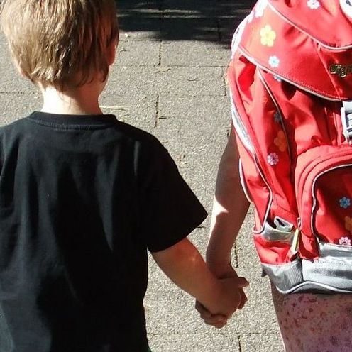 Bildausschnitt von zwei Kinder (Ansicht von hinten), die sich an der hand halten. das rechte tr&auml;gt einen Schulranzen.