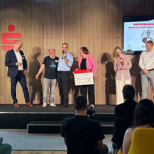 Bas Bild zeigt zwei Frauen und vier M&auml;nner stehen auf einer B&uuml;hne, eine Frau h&auml;lt einen symbolischen Scheck und ein Mann ein Mikrofon.