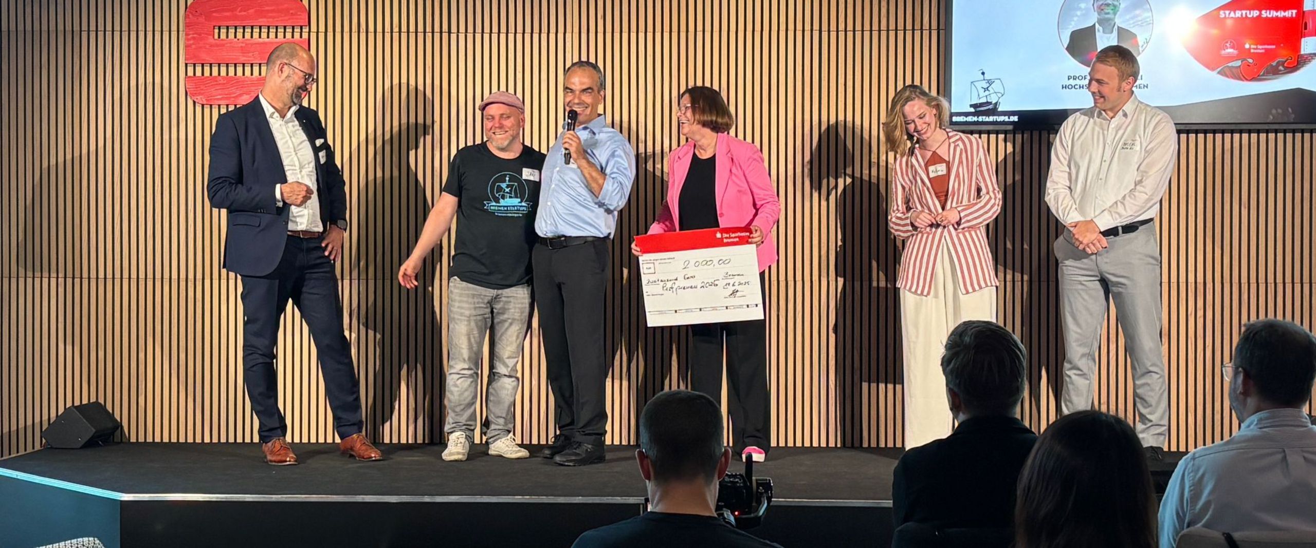 Bas Bild zeigt zwei Frauen und vier M&auml;nner stehen auf einer B&uuml;hne, eine Frau h&auml;lt einen symbolischen Scheck und ein Mann ein Mikrofon.