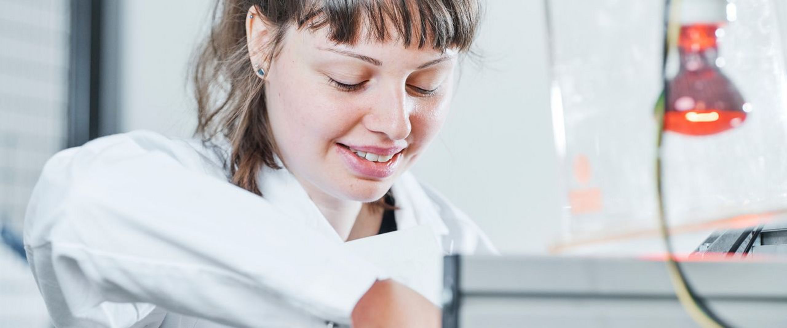 Junge Frau mit Zopf und weißem Kittel schaut runter auf ein Gerät, an dem sie arbeitet.