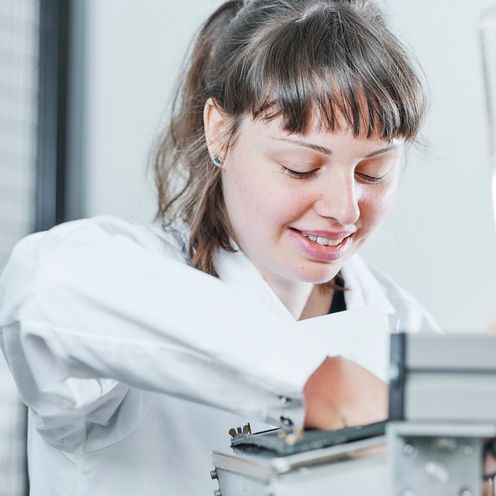Junge Frau mit Zopf und wei&szlig;em Kittel schaut runter auf ein Ger&auml;t, an dem sie arbeitet.