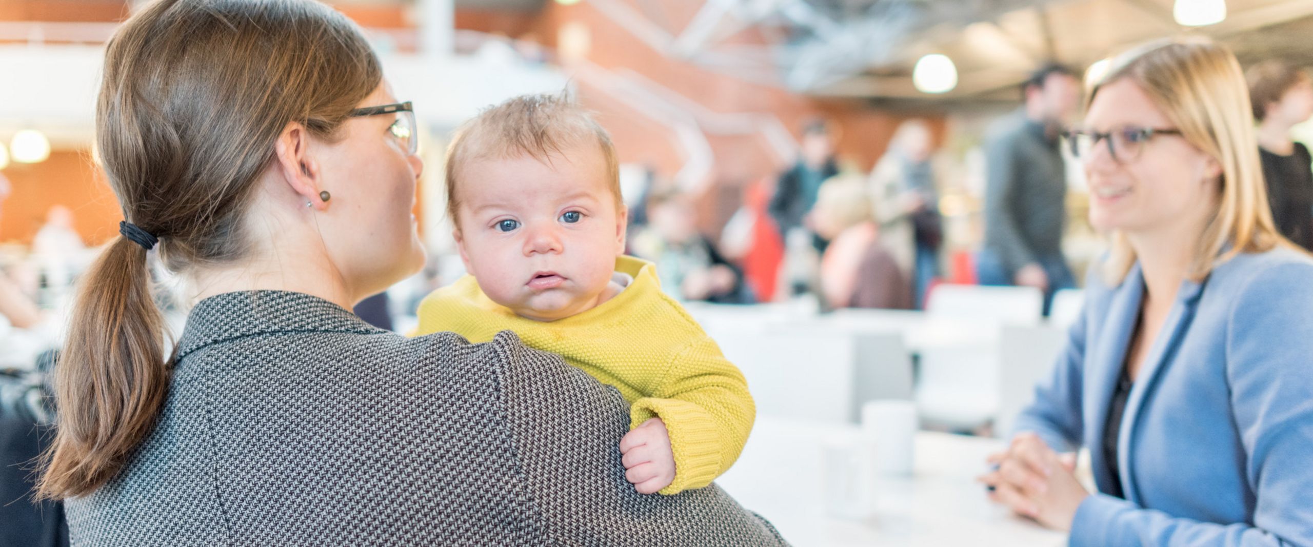 Familienfreundliche Hochschule Eine Professorin sitzt mit ihrem Kleinkind auf dem Arm in der Mensa und unterhält sich mit einer Kollegin.