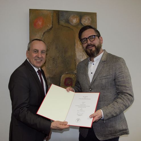 Zwei M&auml;nner halten eine Urkunde und l&auml;cheln in die Kamera