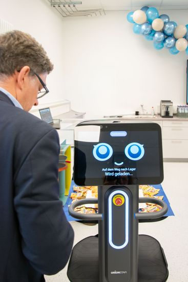 Roboter mit digitalem Gesicht auf einem Tablet schaut in die Kamera. Neben ihm von hinten ein Mann, der zu ihm schaut.