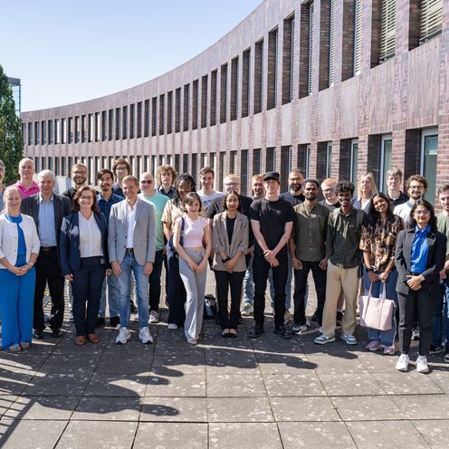 Auf dem Foto sind rund 30 Teilnehmende zusehen. Sie stehen in einer Gruppe zusammen auf einer Terrasse. Im Hintergrund ist ein dunkelrotes Backsteingebäude zu erkennen. Die Sonne scheint. 