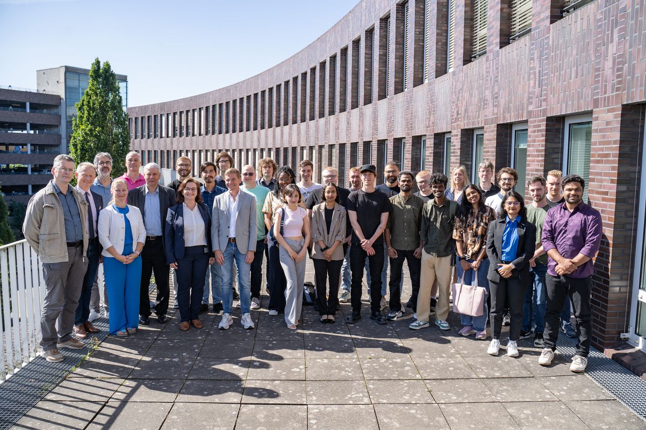 Teilnehmende der EMSS Industry Week 2025 Auf dem Foto sind rund 30 Teilnehmende zusehen. Sie stehen in einer Gruppe zusammen auf einer Terrasse. Im Hintergrund ist ein dunkelrotes Backsteingebäude zu erkennen. Die Sonne scheint.