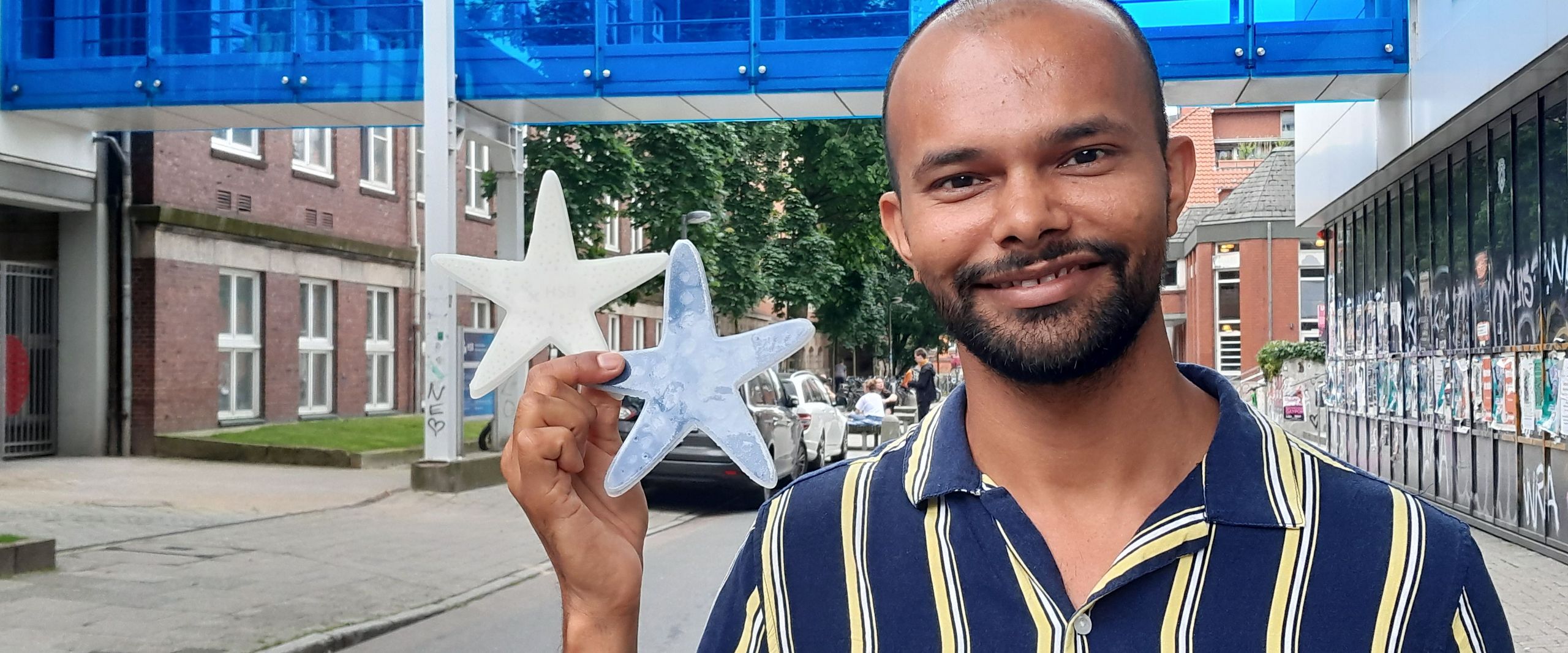 Portrait eines jungen mannes, der in der rechten hand zwei Seesterne aus Silikon hält: einer weiß, einer dunkel. Im Hintergrund sieht man eine blaue Fußgängerbrücke zwischen zwei Gebäuden.