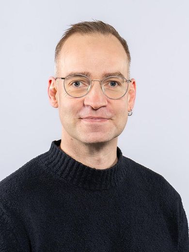 Auf dem Foto ist Roman Balzer zusehen. Er hat kurze blonde Haare. Er tr&auml;gt eine Brille und einen schwarzen Rollkragenpullover. 