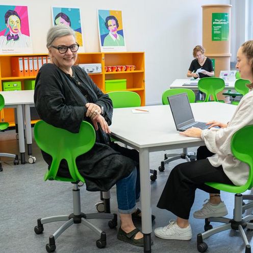 Auf dem Foto ist ein Raum mit bunten M&ouml;beln zusehen. Drei Frauen sitzen verteilt im Raum. Eine Frau schaut direkt in die Kamera. 