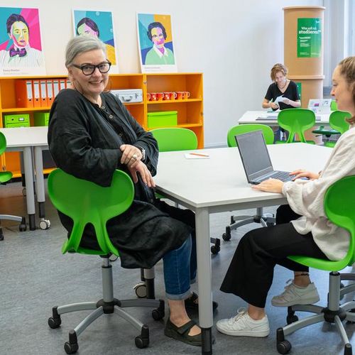Auf dem Foto ist ein Raum mit bunten M&ouml;beln zusehen. Drei Frauen sitzen verteilt im Raum. Eine Frau schaut direkt in die Kamera. 