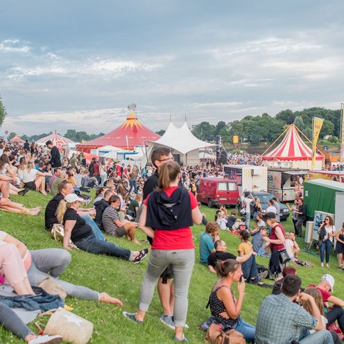 Menschen sitzen bei sommerlichen Temperaturen auf dem Deich. Im Hintergrund sieht man Festivalzelte.