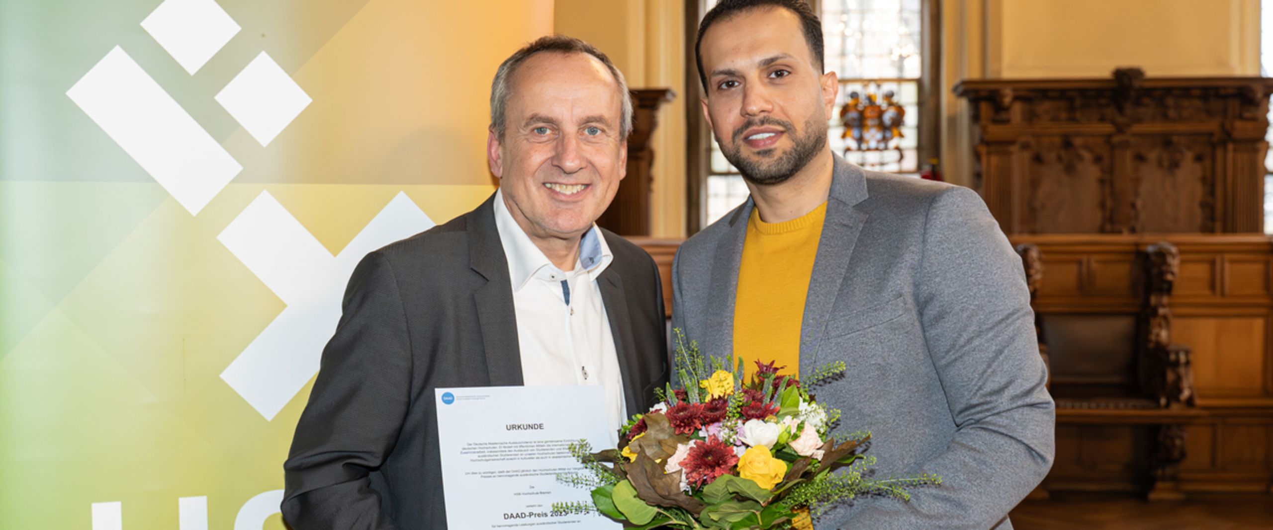 Twei M&auml;nner stehen vor einem banner und schauen in die kamera. Der linke h&auml;lt einen Blumenstrau&szlig;, der rechte eine Urkunde.