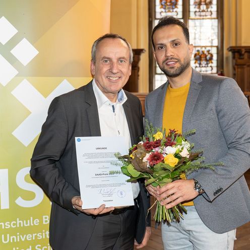 Twei M&auml;nner stehen vor einem banner und schauen in die kamera. Der linke h&auml;lt einen Blumenstrau&szlig;, der rechte eine Urkunde.