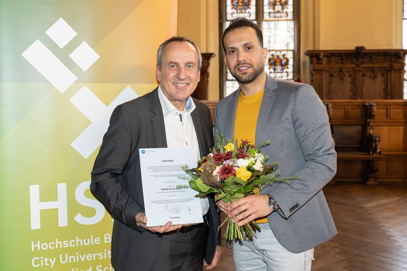 Twei M&auml;nner stehen vor einem banner und schauen in die kamera. Der linke h&auml;lt einen Blumenstrau&szlig;, der rechte eine Urkunde.