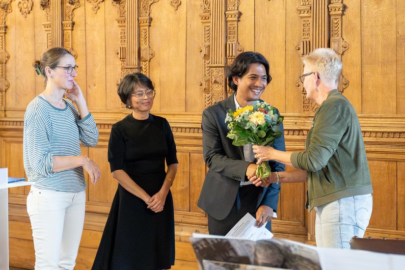 Woman shakes the hand of a young man holding a bouquet of flowers. Two women look on with smiles.