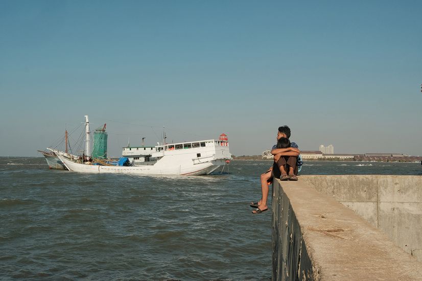 Zwei junge Menschen sitzen auf einer Kaimauer und schauen einem Boot nach, das auf dem Wasser fährt 