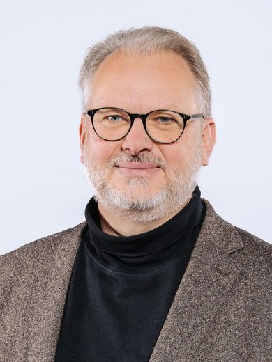 Auf dem Foto ist Michael Pries zusehen. Er hat kurzes graues Haar und einen Bart. Er trägt eine Brille und einen schwarzen Rollkragenpullover unter einem braunen Jackett. 