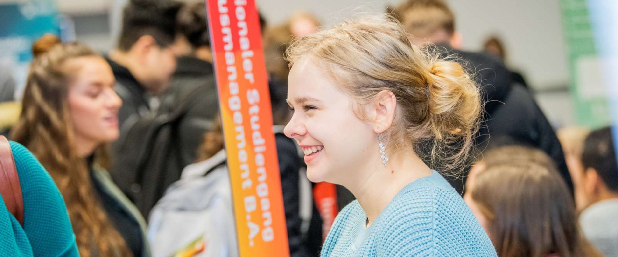 Besucher:innen beim StudienINFOtags. Im Vordergrund und im Fokus ist eine l&auml;chelnde Besucherin zu sehen. Im Hintergrund sind unscharf viele weitere Besucher:innen im Gespr&auml;ch.