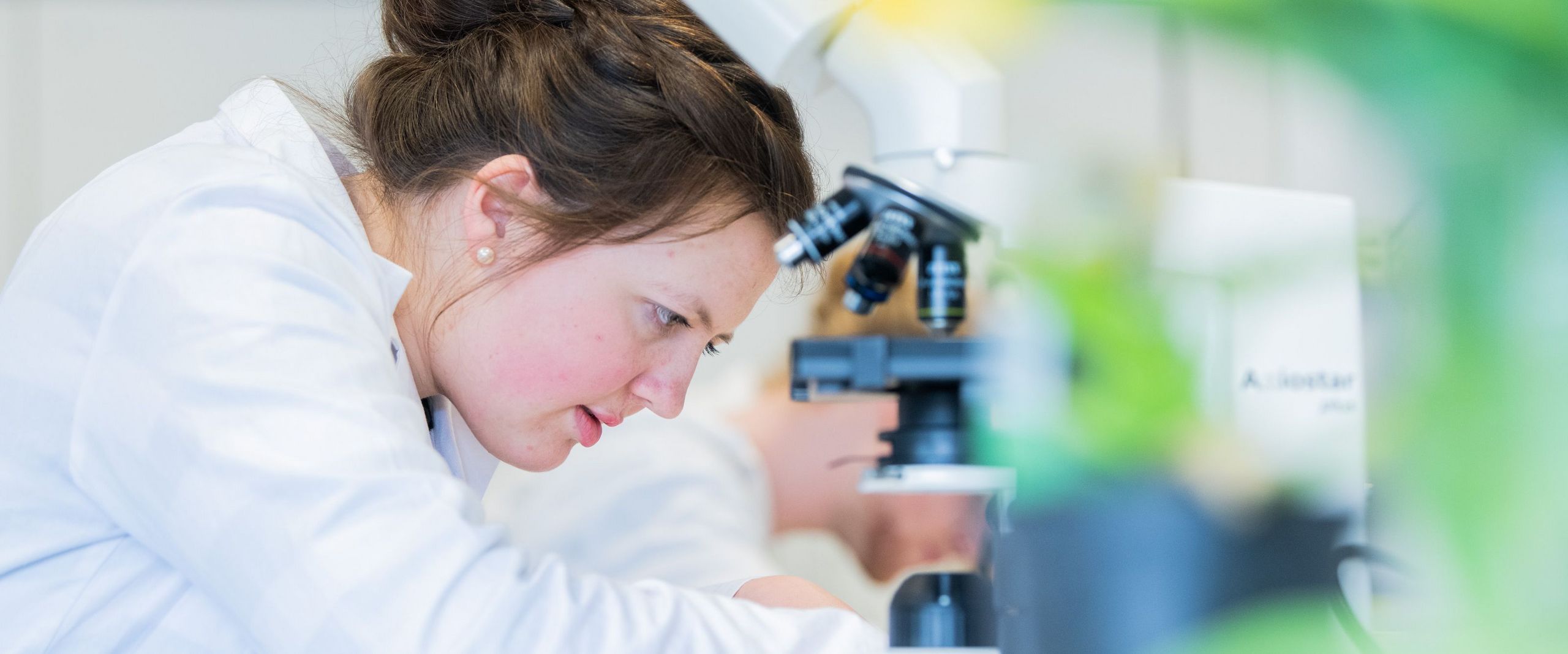 Eine Studentin im Laborkittel beugt sich über ihren Arbeitsplatz neben einem Mikroskop.