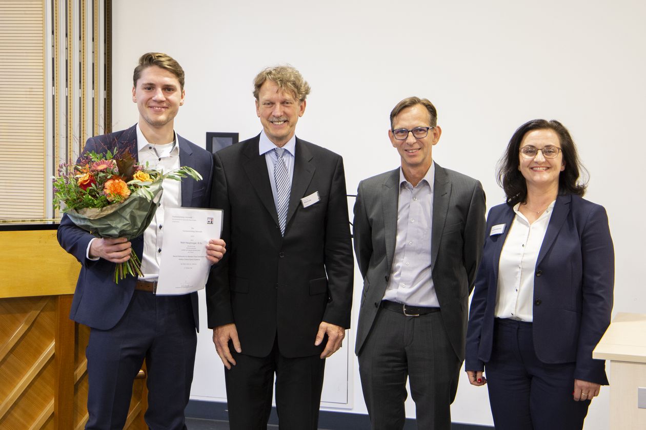 Gruppenbild mit 4 Personen (zwei Männern und einer Frau). Der Mann ganz links hat einen Blumenstrauß in der Hand. Alle lachen in die Kamera.