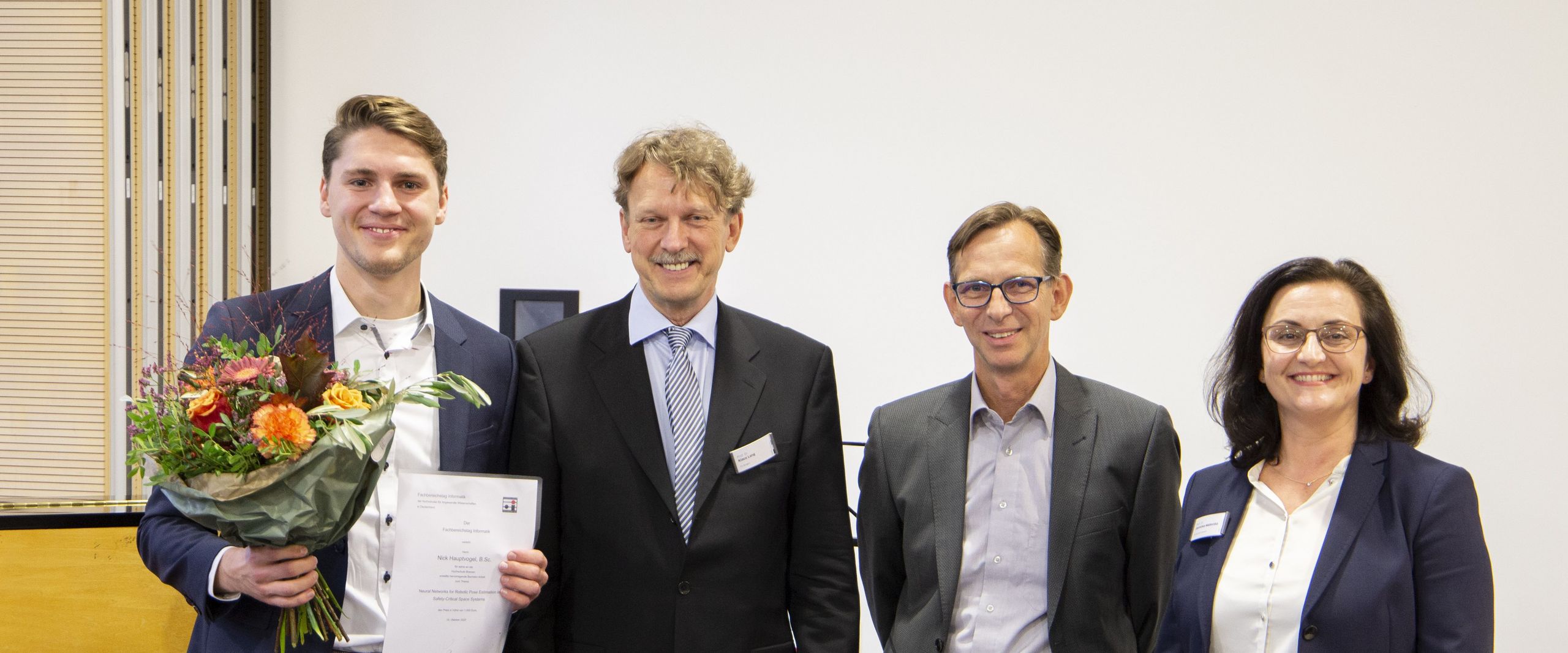 Gruppenbild mit 4 Personen (zwei Männern und einer Frau). Der Mann ganz links hat einen Blumenstrauß in der Hand. Alle lachen in die Kamera.