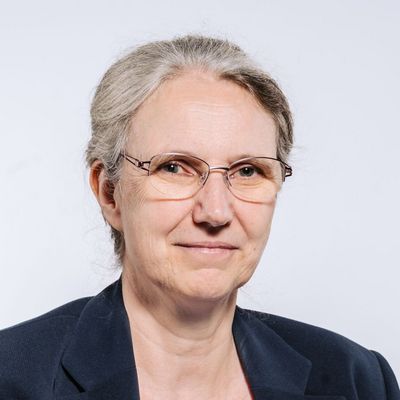 Auf dem Bild ist Martina Harms zusehen. Sie hat ihr graues Haar zu einem lockeren Zopf zurückgeflochten. Sie trägt eine dünne Brille und ein hellblaues T-Shirt unter einem dunkelblauen Blazer.