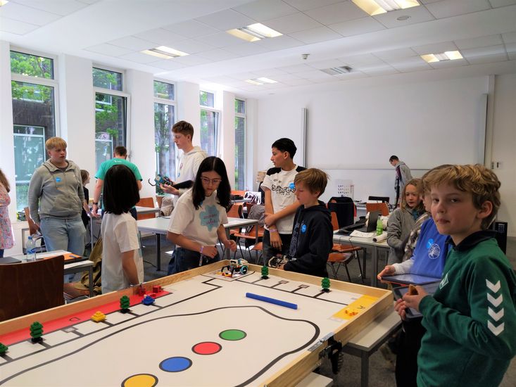 M&auml;dchen und Jungen stehen um ein Spielfeld, auf dem kleine Roboter und Figuren fahren