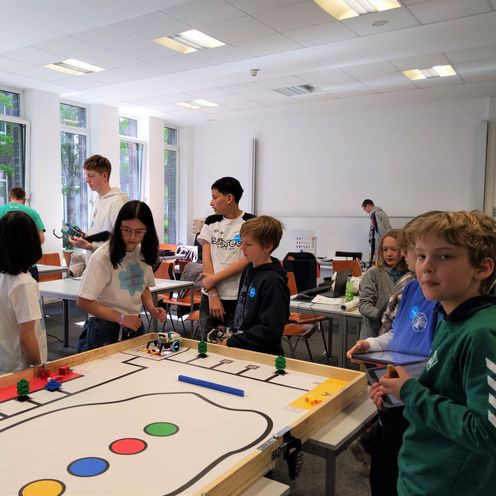 M&auml;dchen und Jungen stehen um ein Spielfeld, auf dem kleine Roboter und Figuren fahren