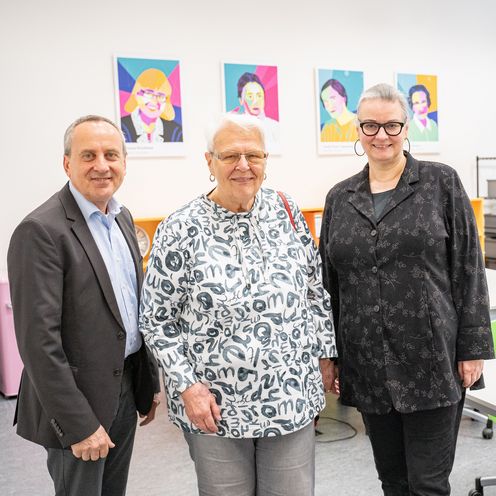 Besuch Brede Stiftung meetMINT-Space gruppenbild mit Mann und zwei Frauen. Im Hintergrund sieht man Tische und bunte Portraits von berühmten MINT-Wissenschaftlerinnen. rechts steht ein grün-weißes Roll-up.