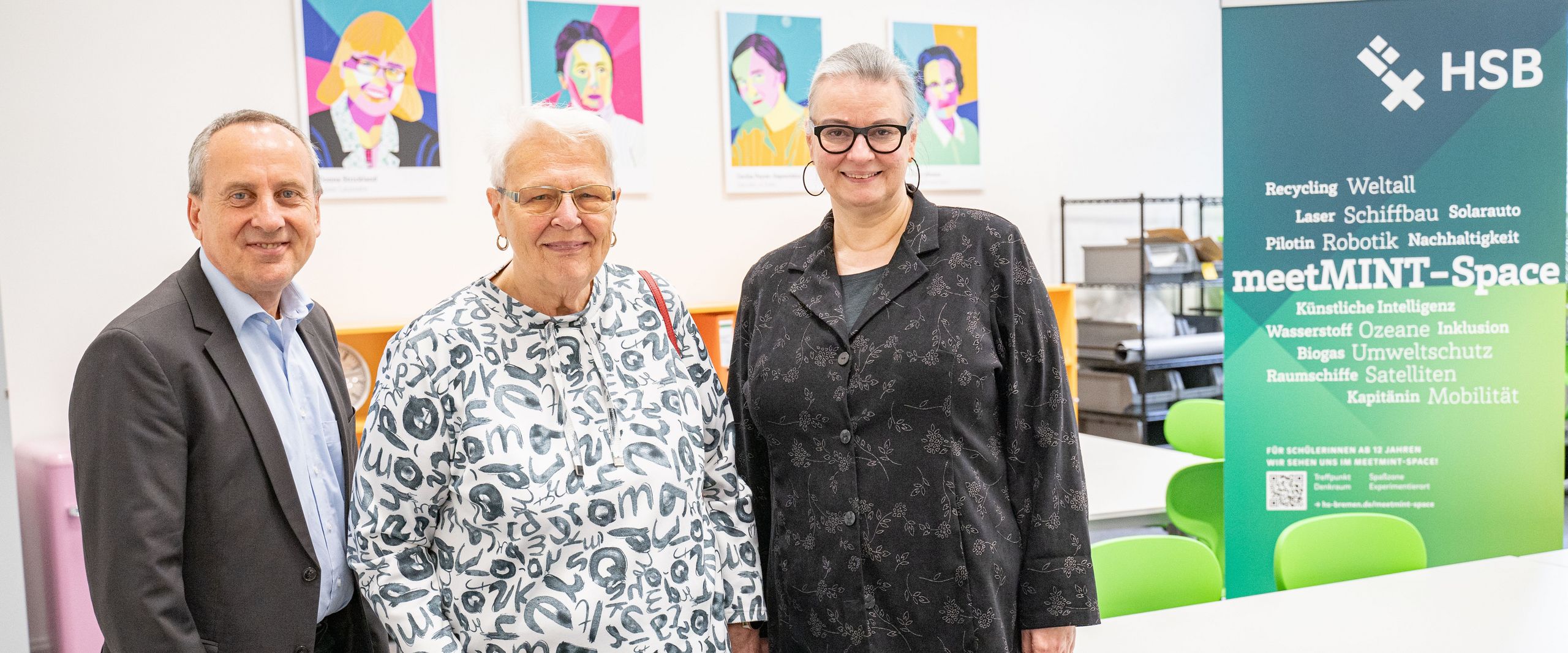 gruppenbild mit Mann und zwei Frauen. Im Hintergrund sieht man Tische und bunte Portraits von berühmten MINT-Wissenschaftlerinnen. rechts steht ein grün-weißes Roll-up.