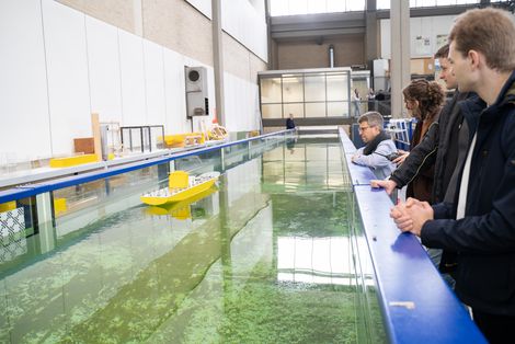 Menschen stehen in einer Werkhalle an einem Wasserkanal und schauen auf ein Modellboot.