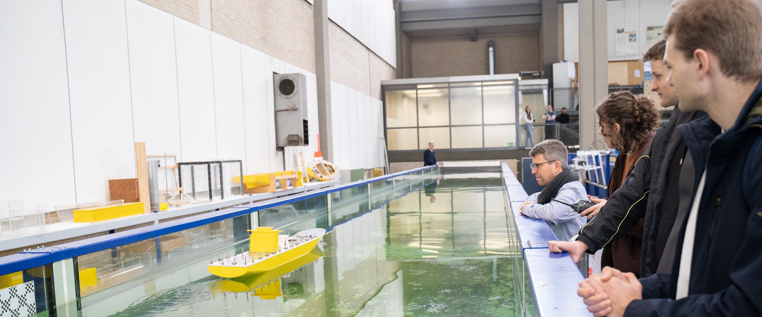 HSB Infotag 2023 Schleppkanal Schiffbau und Meerestechnik Menschen stehen in einer Werkhalle an einem Wasserkanal und schauen auf ein Modellboot.