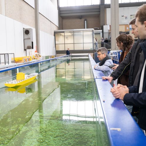 Menschen stehen in einer Werkhalle an einem Wasserkanal und schauen auf ein Modellboot.
