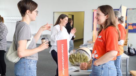 Auf dem Bild sind zwei junge Frauen zu sehen. Die rechte Frau trägt ein rotes T-Shirt der HSB. Sie ist hier Studentin. Die andere Frau trägt ein weiß-schwarz gestreiftes T-Shirt. Sie unterhalten sich. 